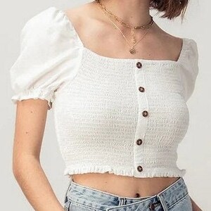 May include: White cropped top with a square neckline, puffed sleeves, and a single button closure. The top has a slightly gathered or smocked design.