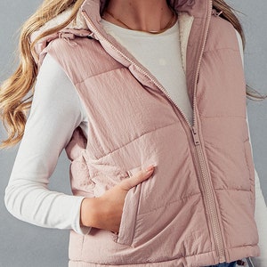 K&ouml;nnte beinhalten: Eine gesteppte Weste in Rosa mit einer Kapuze mit Sherpa-Futter und einem Rei&szlig;verschluss. Die Weste hat eine Tasche vorne und einen Kordelzug am unteren Rand.