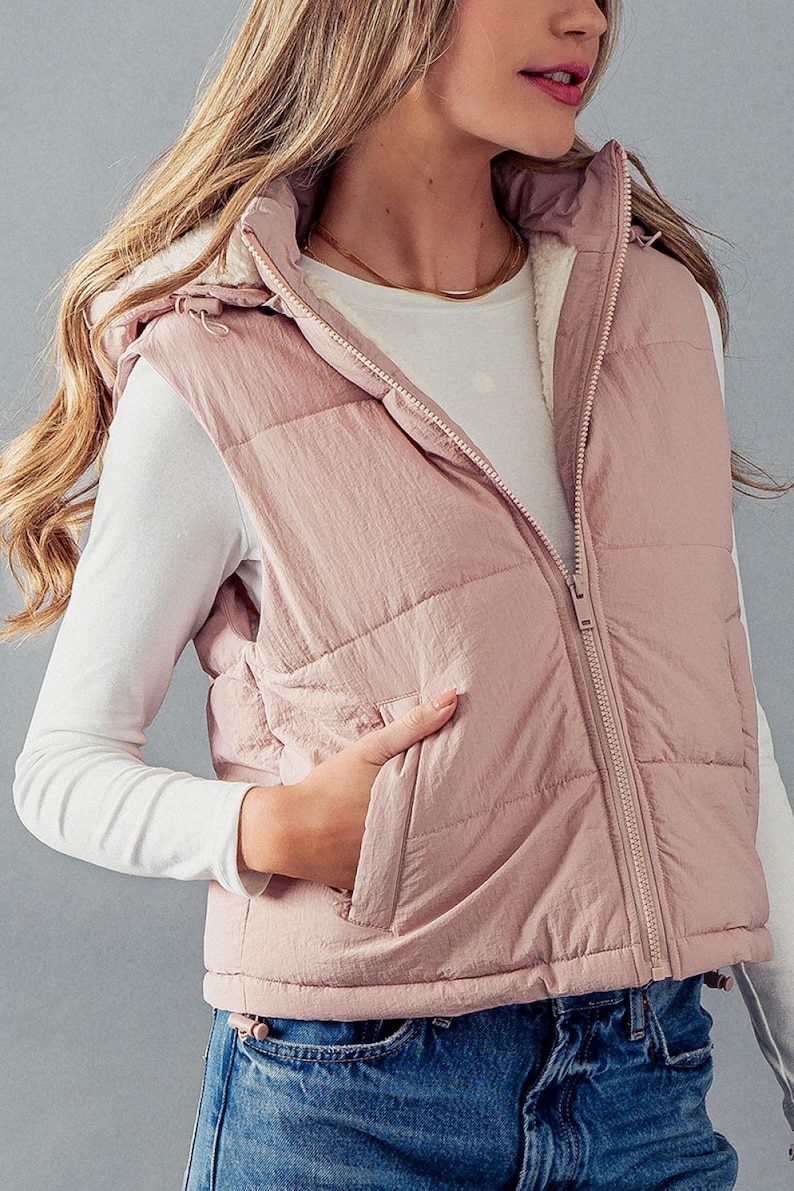 K&ouml;nnte beinhalten: Eine gesteppte Weste in Rosa mit einer Kapuze mit Sherpa-Futter und einem Rei&szlig;verschluss. Die Weste hat eine Tasche vorne und einen Kordelzug am unteren Rand.
