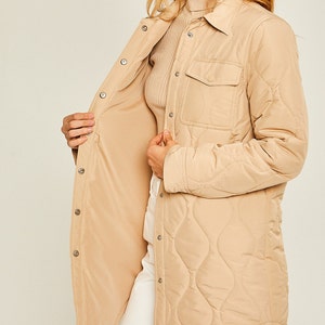 May include: A long-sleeved quilted shirt jacket in a light brown color. The jacket has a button-up front, a chest pocket, and a curved hem.