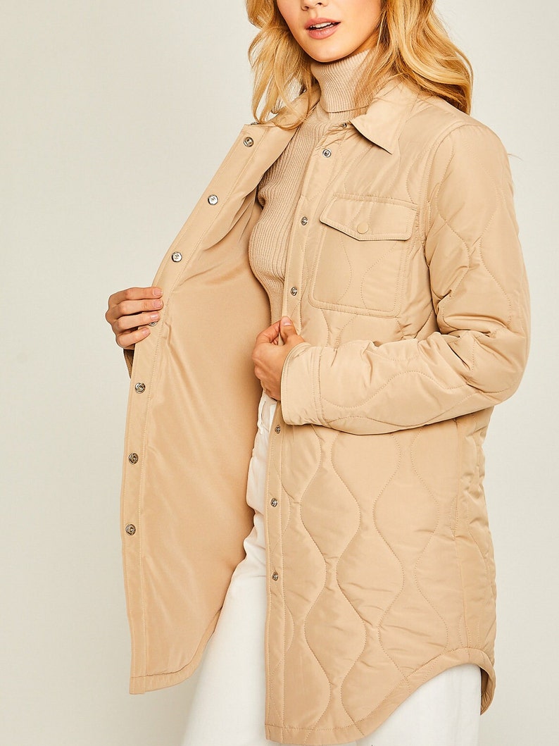 May include: A long-sleeved quilted shirt jacket in a light brown color. The jacket has a button-up front, a chest pocket, and a curved hem.
