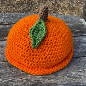 May include: A crocheted orange pumpkin hat with a green leaf and brown stem. The hat is made with a single crochet stitch and has a textured surface.