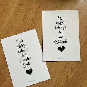 Könnte beinhalten: Zwei weiße Karten mit schwarzem Text und einer schwarzen Herzillustration. Auf einer Karte steht "Mein Herz gehört der dunklen Seite" und auf der anderen "My heart belongs to the darkside". Die Karten liegen auf einer Holzoberfläche.