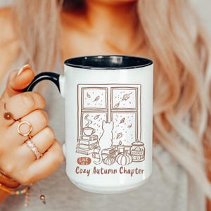 Peut inclure: Mug en céramique blanche avec une anse et un bord noirs. L'illustration marron représente un chat, une fenêtre, des citrouilles et le texte "Lost in a Cozy Autumn Chapter". Un design sur le thème de l'automne.
