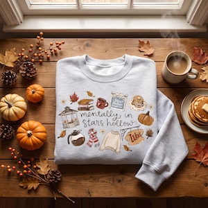 Puede incluir: Una sudadera gris claro de cuello redondo con un estampado gráfico que incluye el texto "mentally in stars hollow" rodeado de ilustraciones de un cenador, libros, una cafetera, una bufanda, un suéter, un letrero de "Luke's" y tortitas.