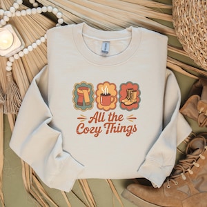 Può includere: Una felpa color crema con la scritta "All the Cozy Things" in un font retrò. Sopra il testo ci sono tre illustrazioni colorate di una sciarpa, una tazza di caffè e stivali. La felpa è un articolo a tema autunnale.