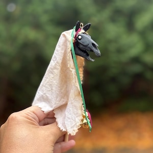 May include: A black horse skull figurine with a cream-colored fabric draped over it. The horse skull has a green and red ribbon with small gold bells. The figurine is held in a hand.