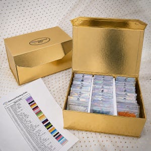 Puede incluir: Una caja de almacenamiento dorada con tapa abatible, que contiene múltiples bolsas de plástico iridiscentes pequeñas llenas de cuentas coloridas para pintura de diamantes. Una caja dorada cerrada y una tabla de colores están cerca.