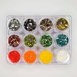 Op de afbeelding: Een doorzichtige plastic container met 12 kleine potjes gevuld met verschillende gekleurde vierkante kralen. De kralen zijn in tinten zilver, goud, rood, groen, blauw, oranje en wit.