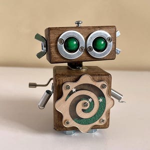 Puede incluir: Un robot de madera hecho a mano con cuerpo y cabeza cuadrados. El robot presenta ojos de cristal verde, detalles metálicos y un panel decorativo de madera con un diseño en espiral. Los brazos del robot son de muelles y varillas metálicas.