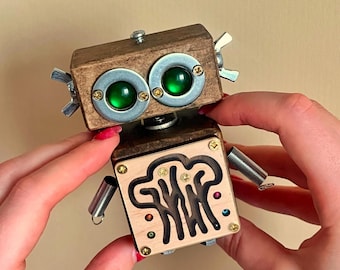 May include: A handcrafted wooden robot toy with a light brown finish. The robot features green glass eyes, metal accents, and a spring-loaded arm. The lower body has a maze design with the word "SWIRL" etched into the wood.