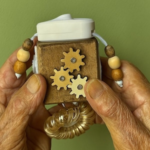Puede incluir: Un cubo sensorial de madera con tres pequeñas piezas de madera en forma de engranaje y una parte superior blanca. El cubo tiene un acabado marrón claro y está adornado con cuentas de madera en cada lado. Una espiral dorada está unida en la parte inferior.