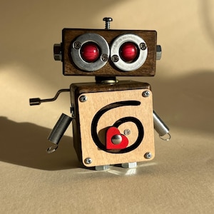 May include: A handcrafted wooden robot sculpture with a square body and rectangular head. The robot features red eye-like accents, metal hardware, and a red heart detail on its front. The arms are made of springs and metal rods.