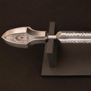 May include: A silver arrow with ornate detailing, including a decorative arrowhead and fletching. The arrow is displayed on a black stand, showcasing its intricate design. The shaft features engraved text.