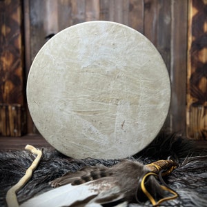 May include: A round, off-white drum with a textured surface, resting on a dark, furry surface. A feathered wand with leather ties and a wooden stick are in the foreground. The background is a wooden wall.
