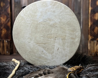 Könnte beinhalten: Eine runde, cremefarbene Trommel mit strukturierter Oberfläche, die auf einer dunklen, pelzigen Oberfläche liegt. Ein Federstab mit Lederbändern und ein Holzstab befinden sich im Vordergrund. Der Hintergrund ist eine Holzwand.