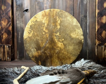 Könnte beinhalten: Eine braune Ledertrommel mit einem naturbelassenen Holzrahmen, die auf einem weißen und grauen Fellteppich liegt. Ein Holzstock und ein Federfächer liegen ebenfalls auf dem Teppich.