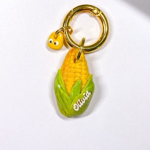 May include: A gold key chain with a yellow and green corn on the cob charm. The charm has the name "Olivia" written on it.