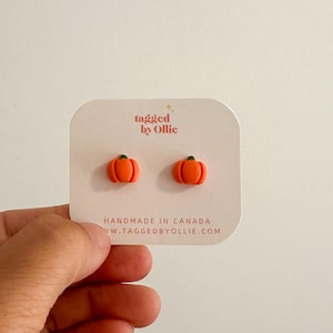 May include: A pair of small orange pumpkin stud earrings with green stems attached to a white display card. The card features the text tagged by Ollie and HANDMADE IN CANADA TAGGEDBYOLLIE.COM printed in a light orange font on the front.