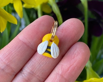 Puede incluir: Un amuleto de oro en forma de abeja con rayas negras y alas blancas. El amuleto está siendo sostenido por una mano contra un fondo de flores amarillas.