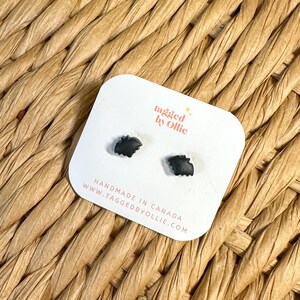 May include: A pair of small black bat-shaped stud earrings attached to a white display card. The card features the text tagged by Ollie, HANDMADE IN CANADA, and the website URL WWW.TAGGEDBYOLLIE.COM printed in black and orange lettering.