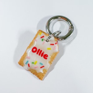 Puede incluir: Llavero rectangular que se asemeja a una pastelería glaseada, con la palabra "Ollie" en rojo sobre el glaseado blanco. La pastelería tiene chispas de colores y está unida a un cierre de metal plateado.