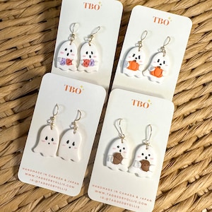 May include: Four pairs of white ghost-shaped dangle earrings on TBO branded display cards. One pair features ghosts with colourful flowers, one with orange pumpkins, one with pink cheeks, and one with brown acorns. Each pair has gold-tone hooks.