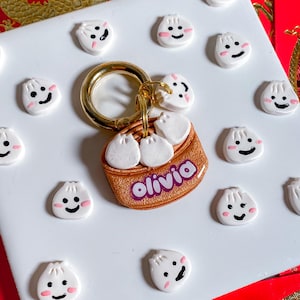 May include: A gold keychain with a brown and gold charm shaped like a steamer basket with three white dumplings inside. The charm has the name "Olivia" written in purple letters. The keychain is surrounded by small white dumplings with pink cheeks and black eyes.