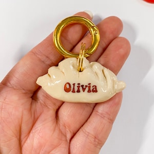 May include: A white ceramic dumpling keychain with a gold metal clasp and the name "Olivia" written in brown lettering.