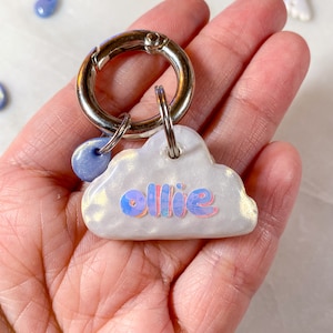 May include: A white cloud-shaped keyring with a silver ring and a blue charm. The cloud has a holographic finish and the name "Ollie" is written in pink and blue letters.