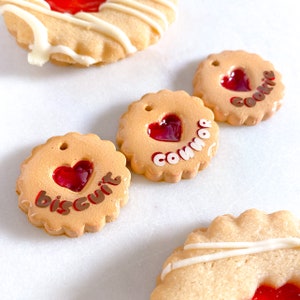 May include: Three decorated sugar cookies with red jelly centres. The cookies are decorated with white icing and the words "Cookie", "Connor", and "Biscuit".