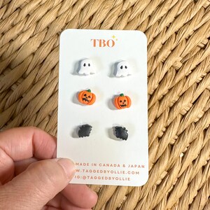 May include: A set of three pairs of stud earrings on a white display card labelled TBO. The earrings feature miniature white ghosts, orange pumpkins with carved faces, and black bats. The card includes the text MADE IN CANADA & JAPAN.