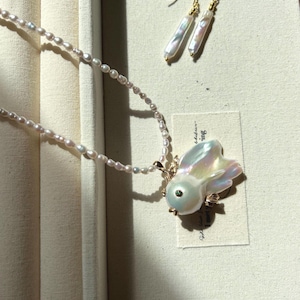 Op de afbeelding: Een witte parelketting met een delicate gouden ketting en een parel en gouden vis hanger. De ketting wordt getoond in een witte doos met een klein kaartje waarop staat "de kleine dingen".