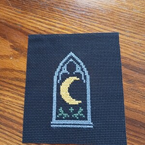 May include: A square piece of black Aida fabric featuring a cross-stitch design of a pointed gothic arch window. Inside the window frame, which is stitched in light blue thread, is a yellow crescent moon above small green leafy sprigs.