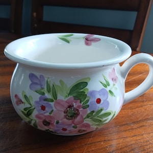 May include: A white ceramic chamber pot featuring a hand-painted floral pattern with pink, purple, and light blue blossoms accented by green leaves. The vessel has a wide, rounded body, a flared rim, and a single curved handle on the right side.