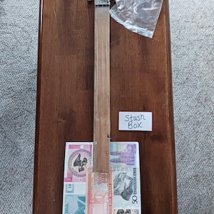 May include: A handmade cigar box guitar with a wooden neck and headstock. The body is decorated with various international banknotes. A small white sign reads "Stash Box". A clear plastic bag and a small metal tube are also present.