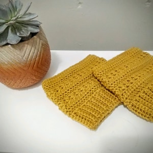 Può includere: Due quadrati di colore giallo senape lavorati a uncinetto, probabilmente sottopentola o sottobicchieri, su una superficie bianca.