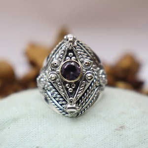 May include: A silver ring with a purple gemstone set in a geometric design. The ring is intricately detailed with twisted wire and bead accents.