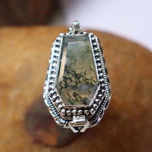 May include: A silver pendant with a green moss agate stone. The pendant is in the shape of a coffin and has a detailed design.