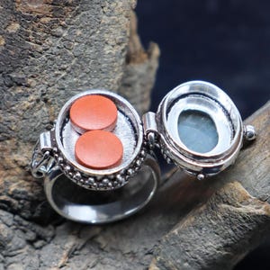 Op de afbeelding: Een zilveren ring met een scharnierend compartiment dat opengaat om twee kleine, ronde, oranje pillen te onthullen. De ring is versierd met een parelmoerontwerp.