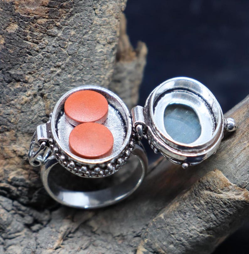 Op de afbeelding: Een zilveren ring met een scharnierend compartiment dat opengaat om twee kleine, ronde, oranje pillen te onthullen. De ring is versierd met een parelmoerontwerp.