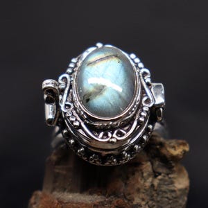 Op de afbeelding: Een zilveren ring met een scharnierend medaillon met een blauwe en groene labradorit edelsteen. Het medaillon heeft ingewikkelde zilveren details en een parelrand.