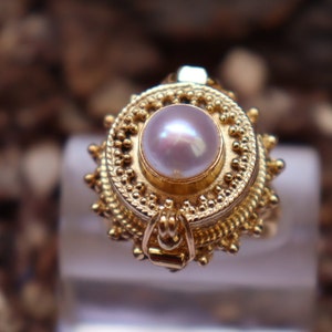 Op de afbeelding: Een gouden ring met een parel in een cirkelvormig ontwerp. De ring heeft een gedetailleerd, sierlijk ontwerp met een scharnierende opening.