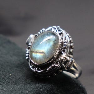 Op de afbeelding: Een zilveren ring met een grote ovale labradorit-edelsteen gezet in een gedetailleerd filigraanontwerp. De ring heeft een scharnierende opening aan de zijkant.