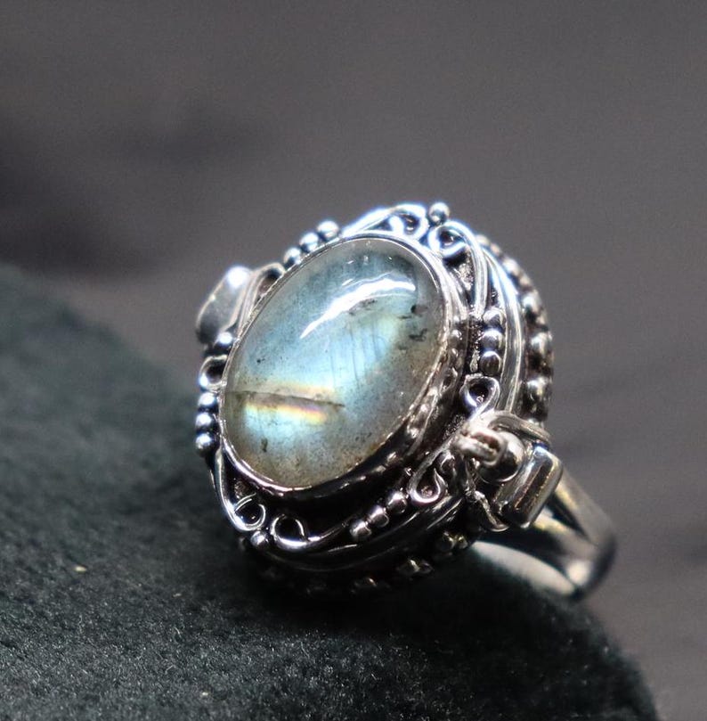 Op de afbeelding: Een zilveren ring met een grote ovale labradorit-edelsteen gezet in een gedetailleerd filigraanontwerp. De ring heeft een scharnierende opening aan de zijkant.