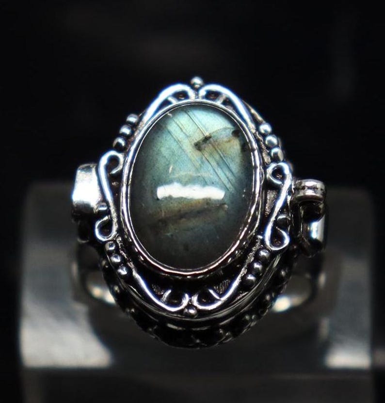 Op de afbeelding: Een zilveren ring met een grote ovale labradorit-edelsteen, gezet in een gedetailleerd filigraanontwerp. De ring heeft een scharnierende opening aan de zijkant.