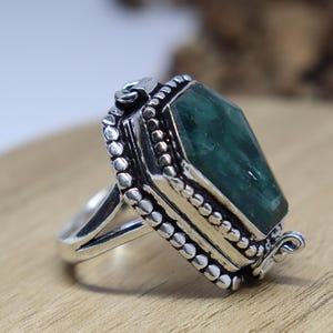 Puede incluir: Un anillo de plata con una gran piedra preciosa verde facetada engastada en un engaste de plata detallado y ornamentado. El anillo tiene una abertura con bisagra en el lateral.