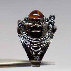 May include: A silver ring with an ornate design and a hinged compartment. The compartment has a round amber stone on top.