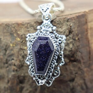 Puede incluir: Un colgante de plata con una piedra azul oscuro brillante en forma de ata&uacute;d. El colgante tiene detalles intrincados y est&aacute; unido a una cadena de plata.