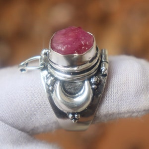 Op de afbeelding: Een zilveren ring met een roze edelsteen in een zilveren zetting. De ring heeft een halve maan ontwerp en een scharnierend deksel dat opent om een klein compartiment te onthullen.